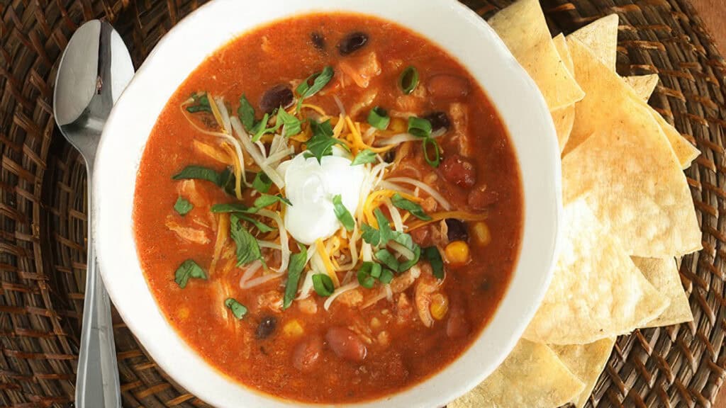 A bowl of hearty taco soup topped with grated cheese, sour cream, and chopped green onions. The soup contains beans, corn, and pieces of chicken in a tomato-based broth. Tortilla chips are served on the side, with a spoon nearby.