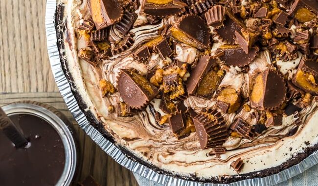 A peanut butter cup pie, the star of all peanut butter desserts, is topped with chunks of cups and a chocolate drizzle. It sits on a wooden table with bowls of whipped cream and extra pieces nearby, plus a jar of chocolate sauce and spoon for that perfect finishing touch.