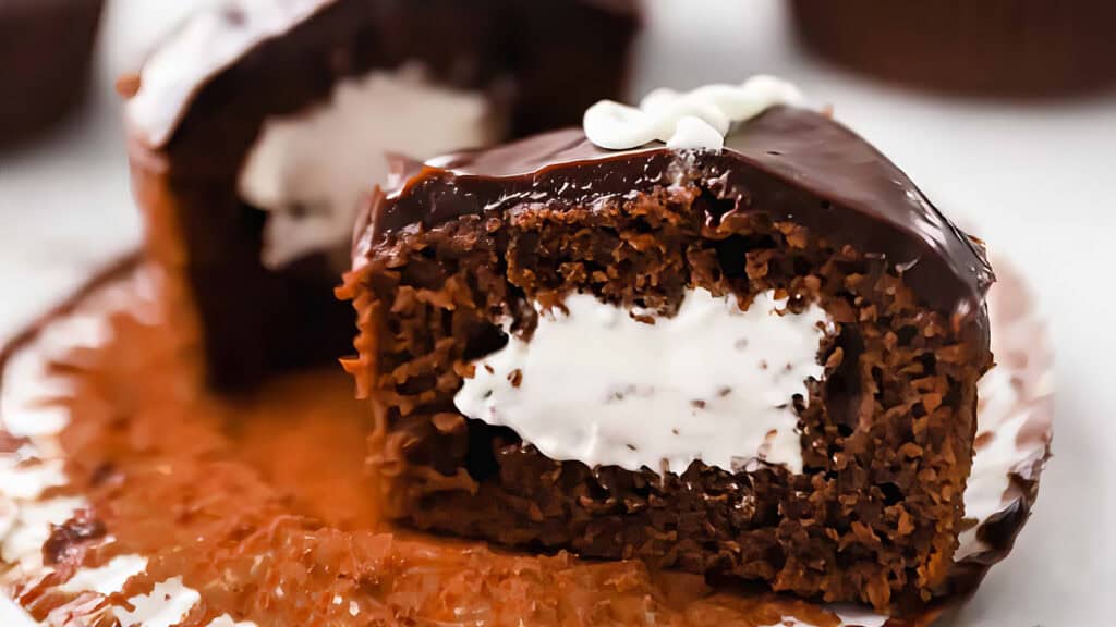 A chocolate cupcake sliced in half reveals a creamy white filling reminiscent of old-fashioned cakes. It's topped with glossy chocolate icing and a small white decorative swirl that keeps everyone coming back for more. It sits on an unwrapped paper liner, inviting indulgence.