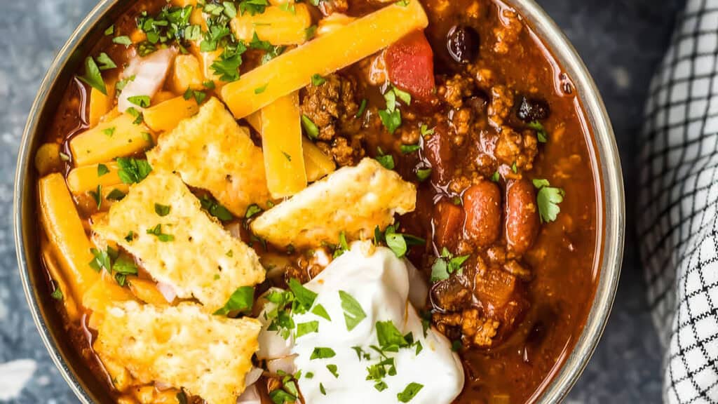 A bowl of slow cooker chili topped with shredded cheese, tortilla chips, sour cream, and fresh herbs. Perfect for cozy nights, this recipe combines beans and ground meat. A checkered napkin lies beside the bowl.