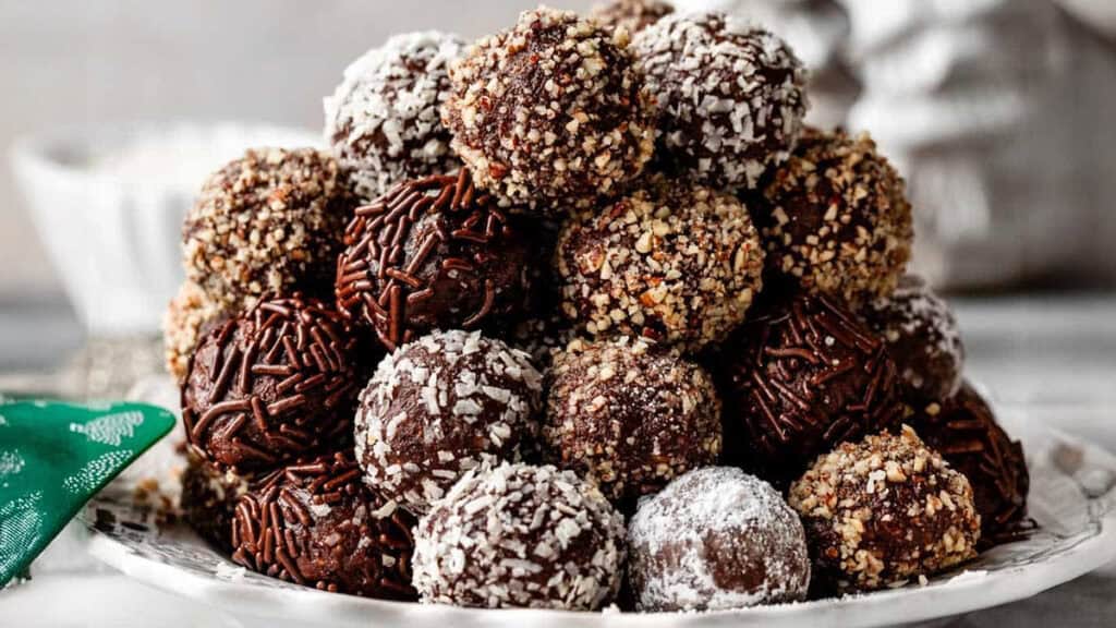 A plate stacked with assorted chocolate truffles, coated in festive toppings like crushed nuts, chocolate sprinkles, powdered sugar, and shredded coconut—perfect holiday treats for any occasion.