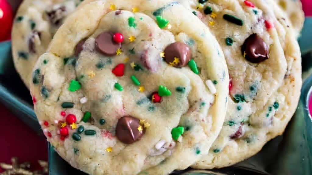 Close-up of colorful cookies topped with chocolate chips and festive sprinkles in shades of red, green, and yellow, arranged on a dark green plate. These delightful treats showcase the magic of holiday baking, boasting a soft, chewy appearance with a joyful holiday theme.