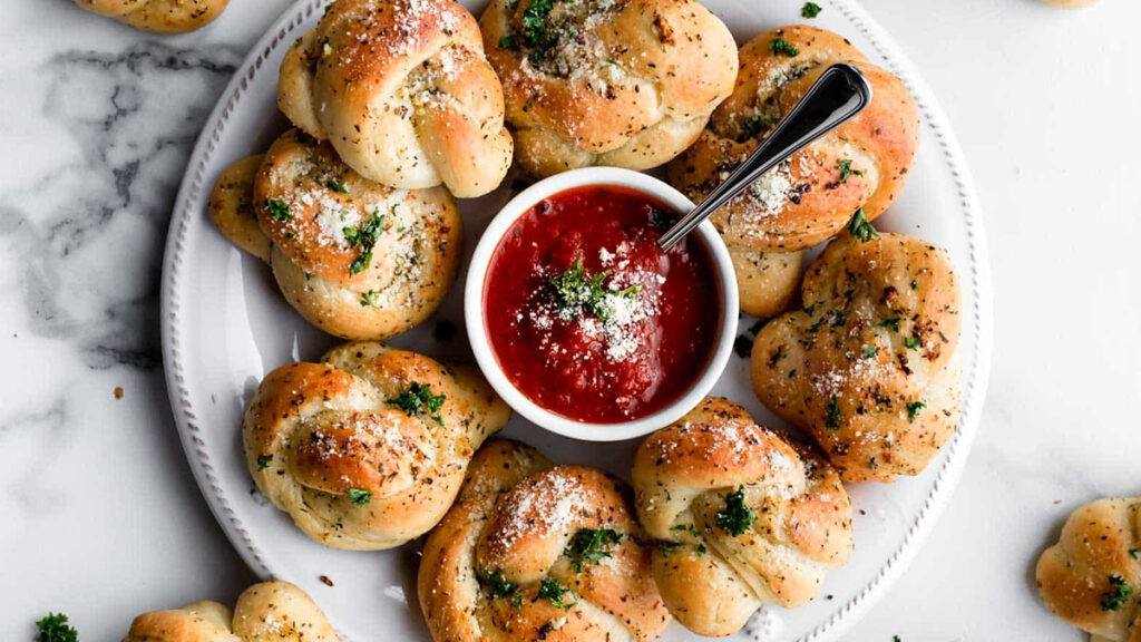 A plate of garlic knots, ideal for finger food recipes, garnished with herbs and Parmesan. They're arranged in a circle around a bowl of red marinara sauce, with a spoon ready to serve, all placed on a marble surface.