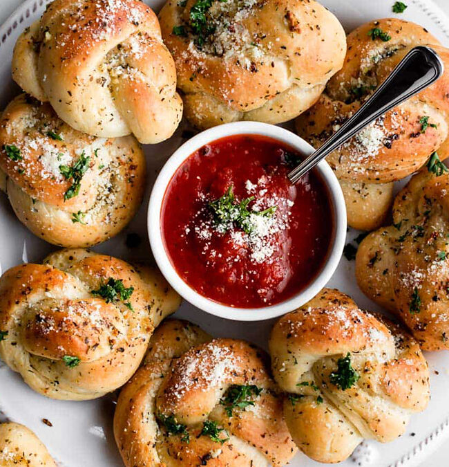 A plate of garlic knots, ideal for finger food recipes, garnished with herbs and Parmesan. They're arranged in a circle around a bowl of red marinara sauce, with a spoon ready to serve, all placed on a marble surface.