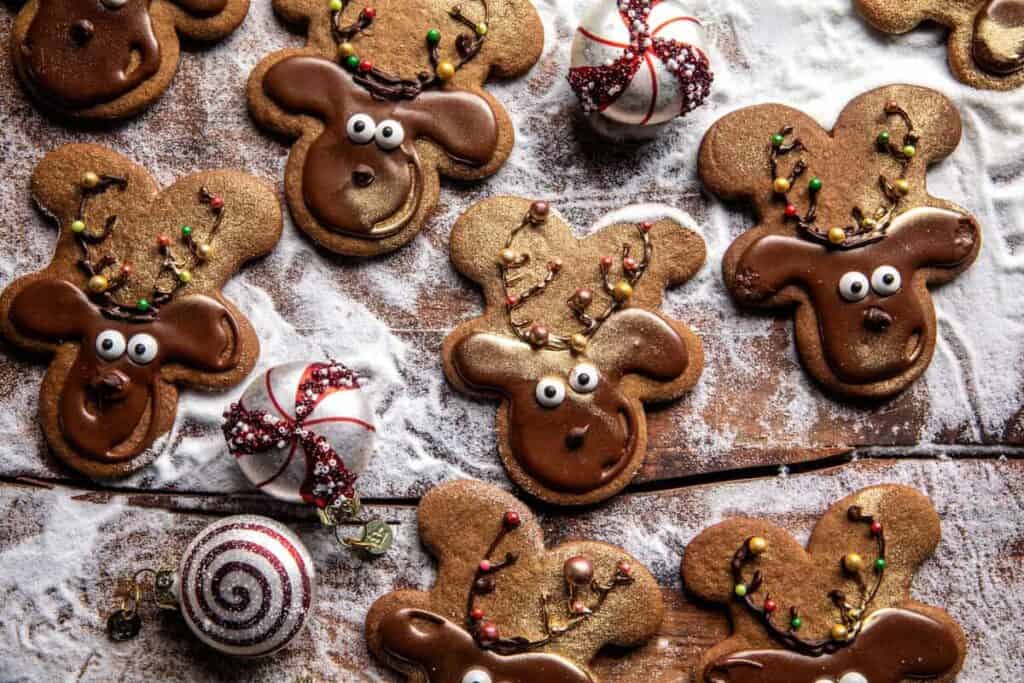 Festive reindeer-shaped cookies with chocolate frosting and candy eyes sit on a wooden surface dusted with powdered sugar, perfect for any holiday baking adventure. They're surrounded by decorative red and white swirled candies, making an irresistible treat from your favorite cookie recipes.