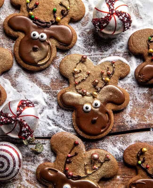 Festive reindeer-shaped cookies with chocolate frosting and candy eyes sit on a wooden surface dusted with powdered sugar, perfect for any holiday baking adventure. They're surrounded by decorative red and white swirled candies, making an irresistible treat from your favorite cookie recipes.