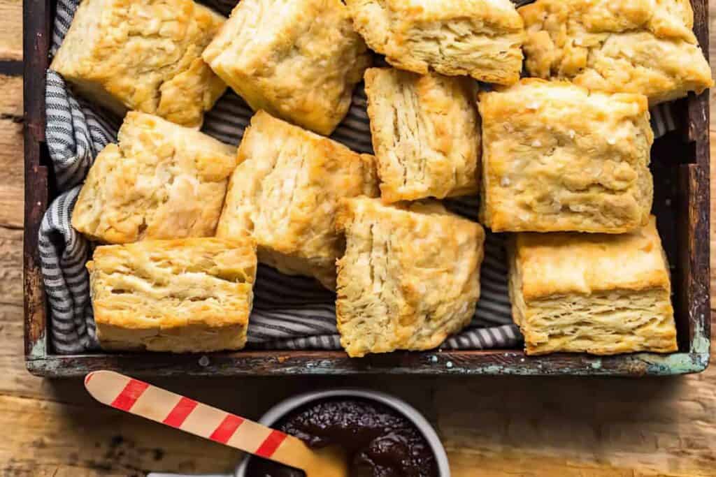 A wooden tray filled with golden, flaky biscuits rests on a striped cloth. Below the tray, a small bowl of dark jam is accompanied by a wooden spreader with a red-striped handle on a rustic wooden surface.