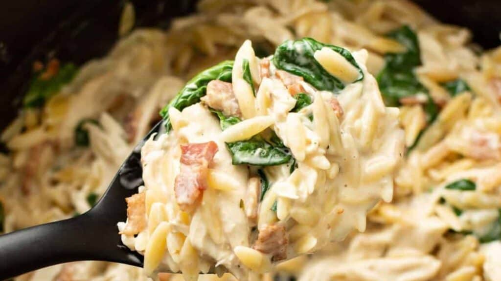 Close-up of a spoon lifting creamy orzo pasta with spinach and bacon from a bowl. This comfort food dish is rich and well-mixed, featuring a smooth, cheesy sauce that lovingly coats the ingredients.