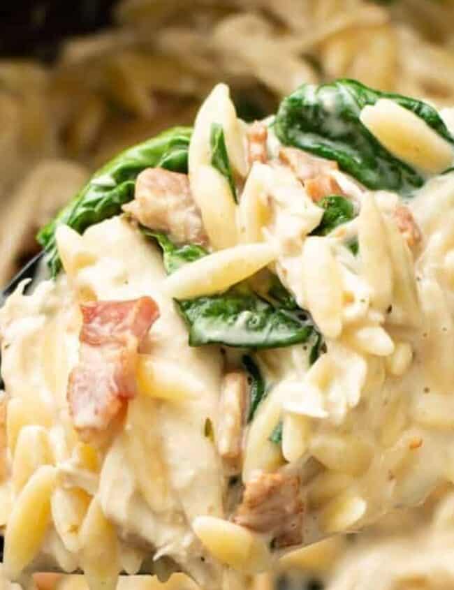 Close-up of a spoon lifting creamy orzo pasta with spinach and bacon from a bowl. This comfort food dish is rich and well-mixed, featuring a smooth, cheesy sauce that lovingly coats the ingredients.