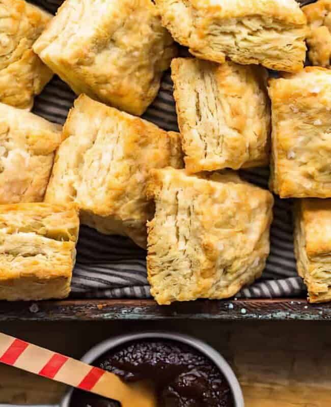 A wooden tray filled with golden, flaky biscuits rests on a striped cloth. Below the tray, a small bowl of dark jam is accompanied by a wooden spreader with a red-striped handle on a rustic wooden surface.