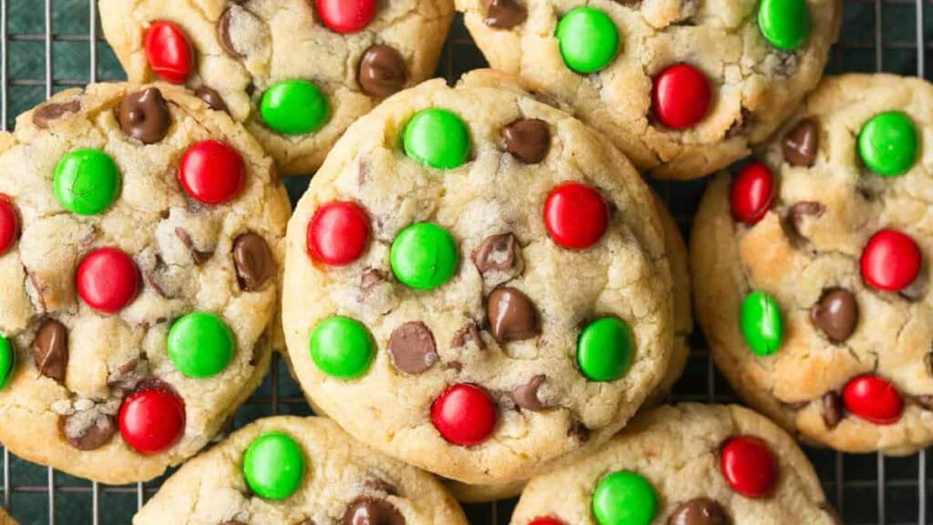 A close-up of holiday treats features cookies topped with red and green candy-coated chocolates and chocolate chips, all beautifully arranged on a cooling rack.