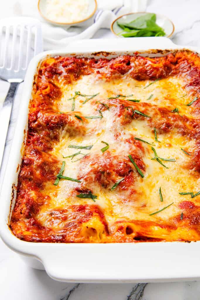 A cheesy baked lasagna in a white dish, topped with melted cheese and garnished with chopped basil. The edges are golden brown and slightly crispy, enhanced by the rich addition of Italian sausage, with a spatula and small bowls in the background on a marble surface.