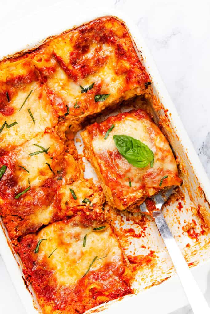 A freshly baked Italian lasagna in a white dish with one slice removed. This cheesy lasagna features a golden, melted cheese topping and is garnished with a fragrant basil leaf.