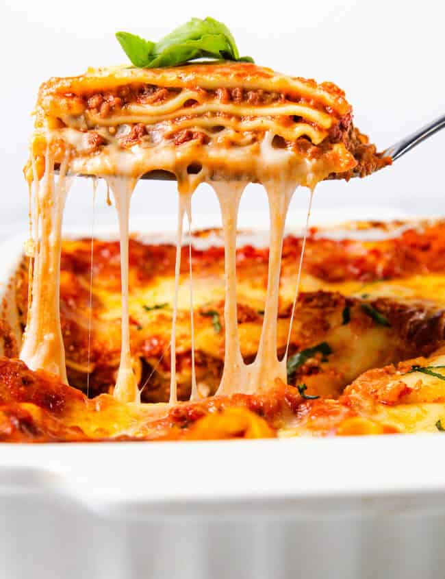 A close-up of a cheesy slice of lasagna being lifted out of a white baking dish. The lasagna features layers of pasta, rich Italian sausage sauce, and melted cheese stretching as it is served. A basil leaf garnishes the top.