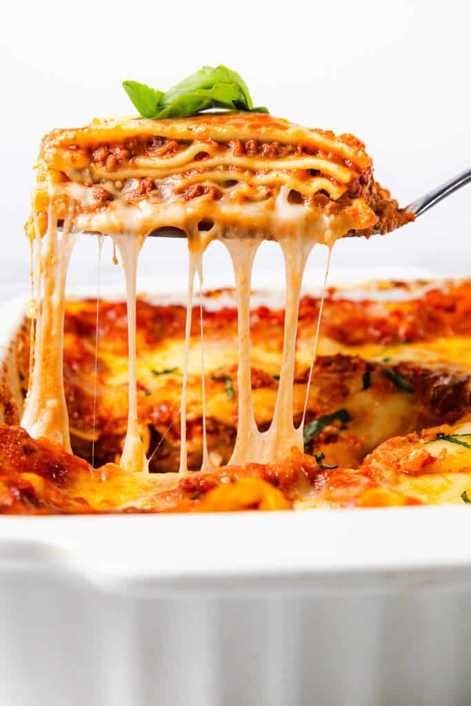 A close-up of a cheesy slice of lasagna being lifted out of a white baking dish. The lasagna features layers of pasta, rich Italian sausage sauce, and melted cheese stretching as it is served. A basil leaf garnishes the top.