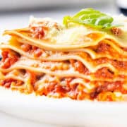 A close-up of a slice of Italian sausage lasagna on a white plate. The cheesy delight boasts multiple layers of pasta, rich meat sauce, and melted cheese, topped with a sprig of fresh basil and garnished with a sprinkle of grated cheese.