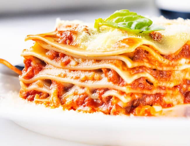 A close-up of a slice of Italian sausage lasagna on a white plate. The cheesy delight boasts multiple layers of pasta, rich meat sauce, and melted cheese, topped with a sprig of fresh basil and garnished with a sprinkle of grated cheese.