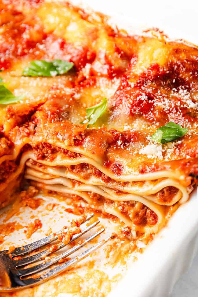 Close-up of a freshly baked, cheesy lasagna in a white dish, with one slice removed. Layers of pasta, Italian sausage meat sauce, and melted cheese are visible. Garnished with fresh basil leaves and a sprinkle of grated cheese, with a fork resting beside it.