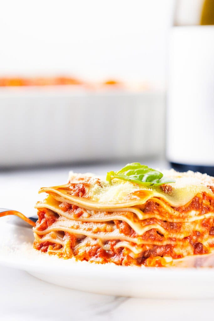 A close-up of a cheesy lasagna slice showcases visible layers of pasta, rich tomato sauce, and melted cheese. Garnished with a basil leaf on a white plate, it's irresistible. In the background, a blurred casserole dish with Italian sausage hints at more deliciousness to come.