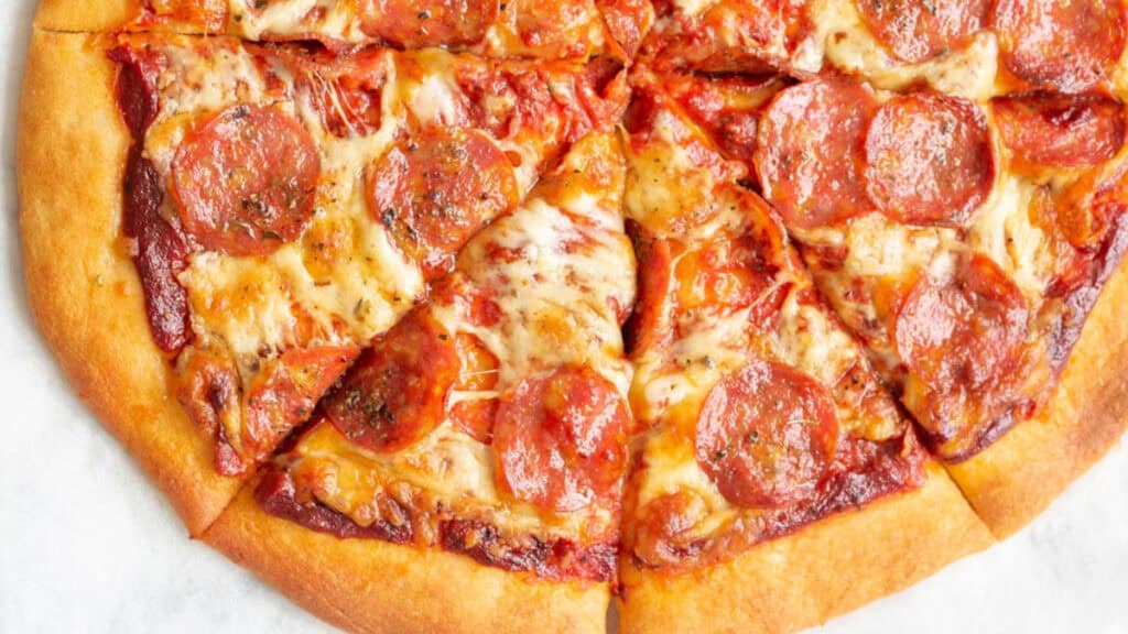 A cooked pepperoni pizza cut into slices with a golden-brown crust, topped with melted cheese and slices of pepperoni, shown from above on a light surface.