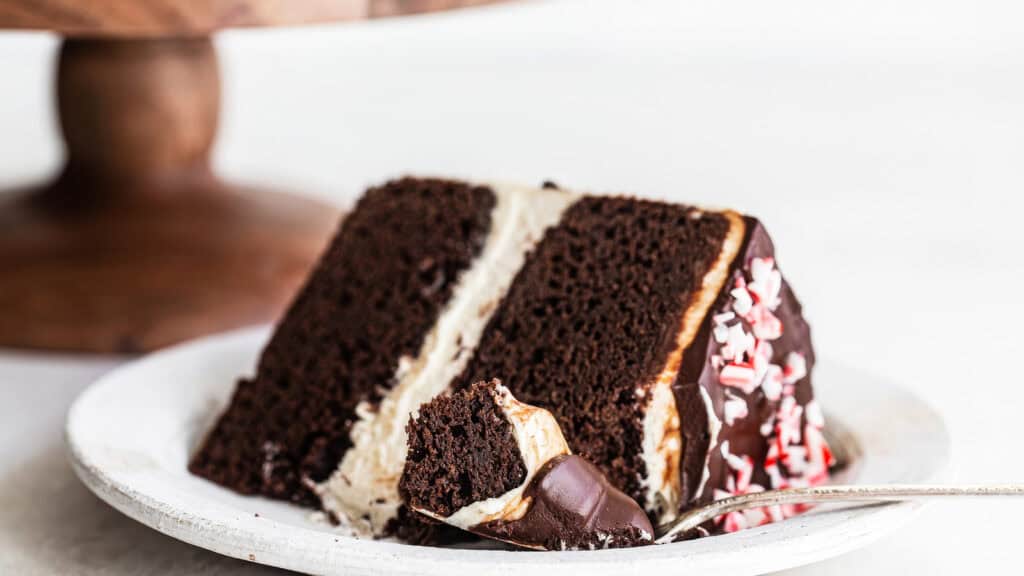 A close-up of a slice from a chocolate cake recipe on a white plate showcases its two layers with white frosting in between. The top gleams with chocolate glaze and sprinkles, while a fork with a small bite rests invitingly beside the dessert.