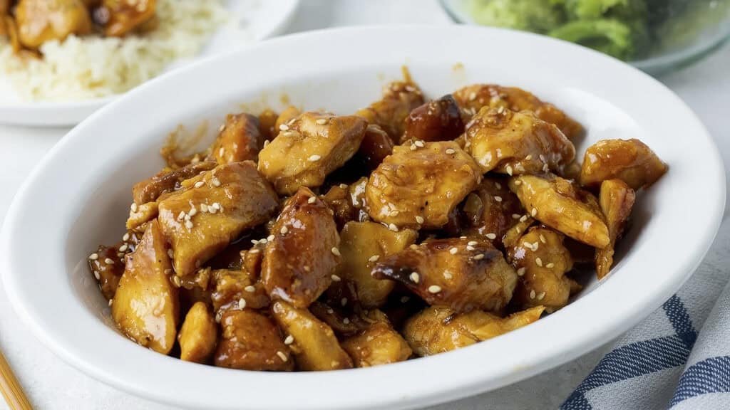 A white oval dish brimming with slow cooker teriyaki chicken chunks, garnished with sesame seeds. The dish sits on a table beside a bowl of rice, with a hint of broccoli in the background—a delicious addition to your collection of slow cooker recipes.