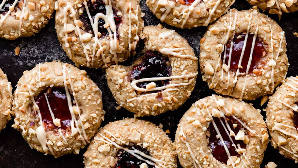 A close-up of several thumbprint cookies arranged on a dark surface. Each cookie recipe features a topping of crushed nuts and a drizzle of white icing. The center is filled with glossy red jam, making these thumbprint cookies irresistible delights.