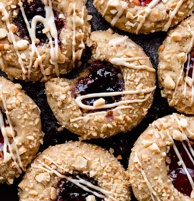 A close-up of several thumbprint cookies arranged on a dark surface. Each cookie recipe features a topping of crushed nuts and a drizzle of white icing. The center is filled with glossy red jam, making these thumbprint cookies irresistible delights.