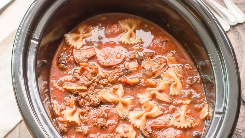 A slow cooker containing a hearty stew with bow-tie pasta, sliced carrots, ground beef, and beans in a rich tomato-based sauce offers a delightful twist on traditional recipes.