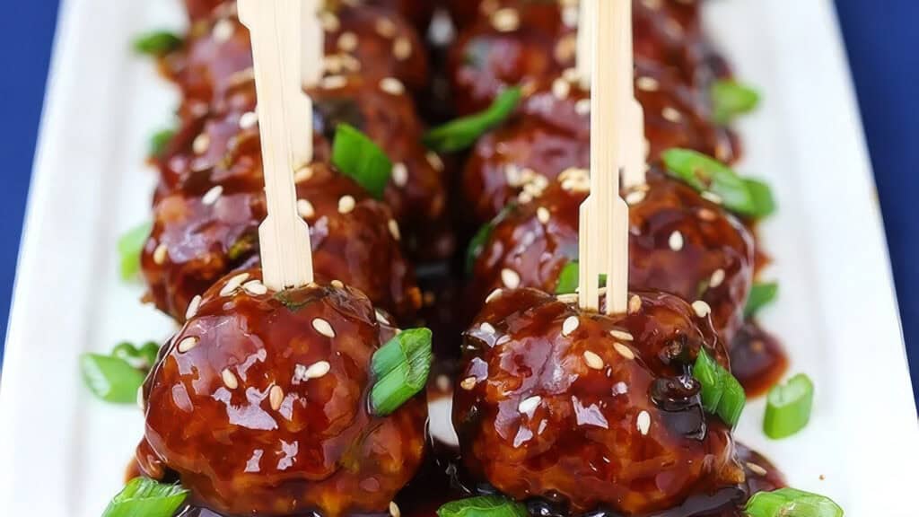 Close-up of glazed meatballs made from flavorful ground beef recipes, accented with sesame seeds and chopped green onions. They're skewered on wooden sticks and artfully arranged on a white rectangular plate. The glossy sauce enhances their savory appeal.