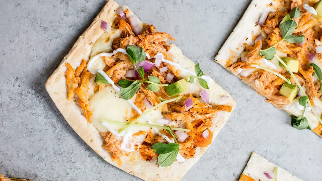 A slice of flatbread topped with shredded chicken, melted cheese, sliced red onion, avocado, greens, and a drizzle of sauce creates a delightful and nutritious option for those seeking skinny recipes. Enjoy this guilt-free indulgence on a gray surface.