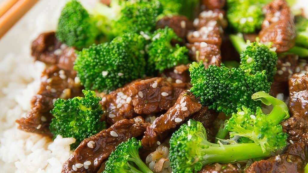 A close-up of a dish featuring tender beef strips mixed with vibrant green broccoli. The dish is garnished with sesame seeds and served over a bed of white rice. Chopsticks are visible in the top corner, suggesting an Asian-inspired meal.