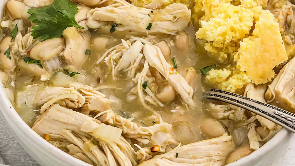 A bowl of white chicken chili topped with cilantro, featuring shredded chicken, white beans, and spices in a broth. Cornbread is crumbled on the side, and a spoon rests in the bowl.