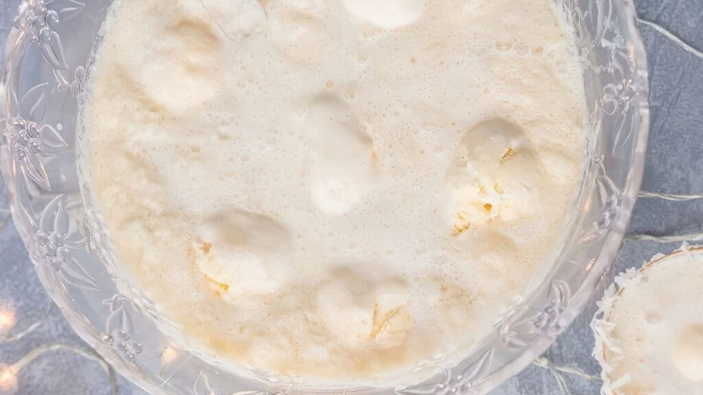 A close-up of a glass bowl filled with a frothy beverage topped with dollops of creamy whipped topping, resembling a punch or dessert. The edges of the bowl feature an ornate, decorative design.