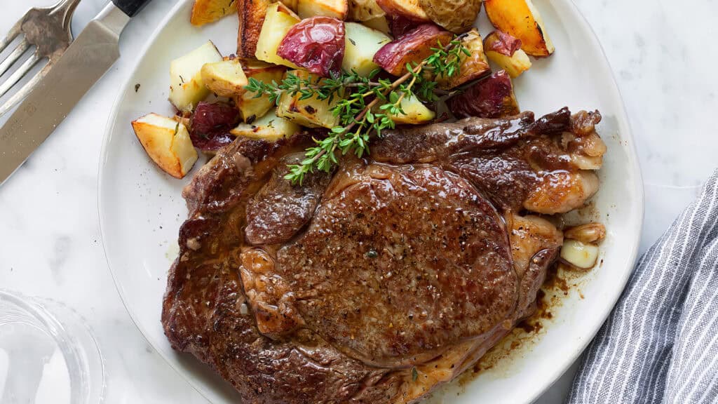 A cooked ribeye steak seasoned with pepper and salt, garnished with fresh thyme, takes center stage among main dishes on a marble surface. It’s served alongside crispy roasted potatoes and evokes the festive spirit of Christmas. A fork and knife await eagerly next to the plate.