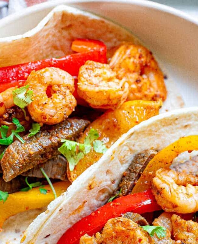 Two flour tortillas filled with shrimp, sliced beef, and vibrant bell peppers make for delicious dinners. Garnished with fresh herbs and served on a white plate, this dish is perfect for weeknight meals. Nearby, a fork and napkin await to complement the fast dining experience.