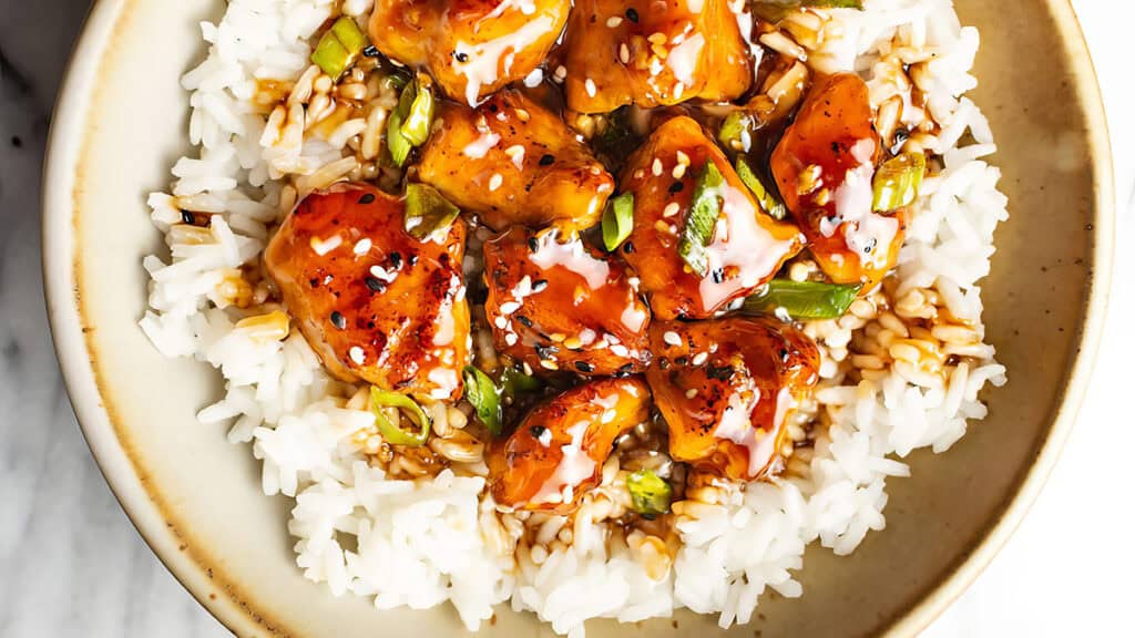 A bowl of white rice topped with glazed chicken pieces and sliced green onions, sprinkled with sesame seeds. Perfect for easy chicken recipes, this dish is presented in a beige bowl on a light surface.