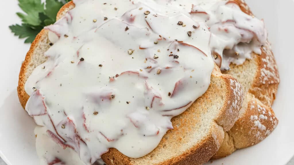 A plate of old-fashioned comfort food: toast topped with creamy white sauce and slices of chipped beef. The dish is garnished with cracked black pepper and a parsley leaf.