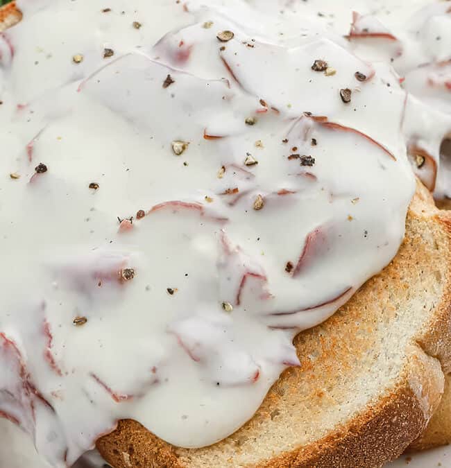 A plate of old-fashioned comfort food: toast topped with creamy white sauce and slices of chipped beef. The dish is garnished with cracked black pepper and a parsley leaf.