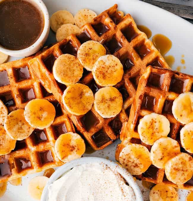 A plate of golden brown waffles topped with banana slices, showcasing one of the finest banana recipes, drizzled with syrup. A small dish of syrup and a dollop of whipped cream garnish the plate. In the background, a fork and a blue napkin complete the inviting scene.