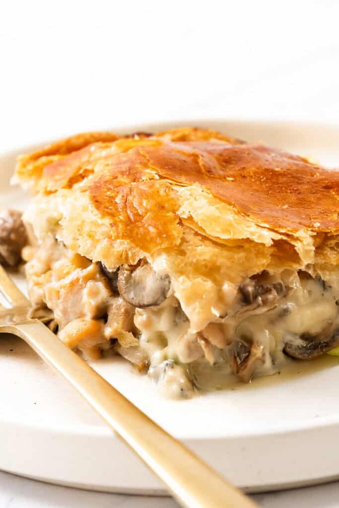 A slice of cheesy chicken and mushroom pie with a creamy filling encased in a golden, flaky crust rests on a white plate, accompanied by a gold fork. The pie reveals chunks of chicken and mushrooms enveloped in a rich sauce.