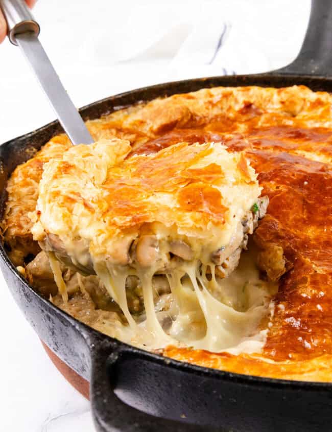 A skillet filled with a Cheesy Chicken & Mushroom Pie is shown. A large slice is being lifted, showcasing a golden, flaky crust and creamy filling with visible chicken, mushrooms, and vegetables. Melted cheese stretches from the main pie to the slice being served.