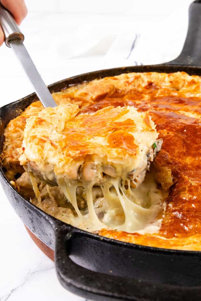 A skillet filled with a Cheesy Chicken & Mushroom Pie is shown. A large slice is being lifted, showcasing a golden, flaky crust and creamy filling with visible chicken, mushrooms, and vegetables. Melted cheese stretches from the main pie to the slice being served.