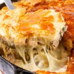 A close-up of a golden brown Cheesy Chicken & Mushroom Pie being served from a skillet. The pie has a flaky crust, and a portion reveals a rich, creamy filling with melted cheese and chunks of chicken enveloping savory mushrooms.