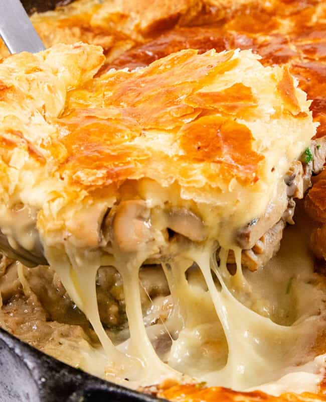 A close-up of a golden brown Cheesy Chicken & Mushroom Pie being served from a skillet. The pie has a flaky crust, and a portion reveals a rich, creamy filling with melted cheese and chunks of chicken enveloping savory mushrooms.