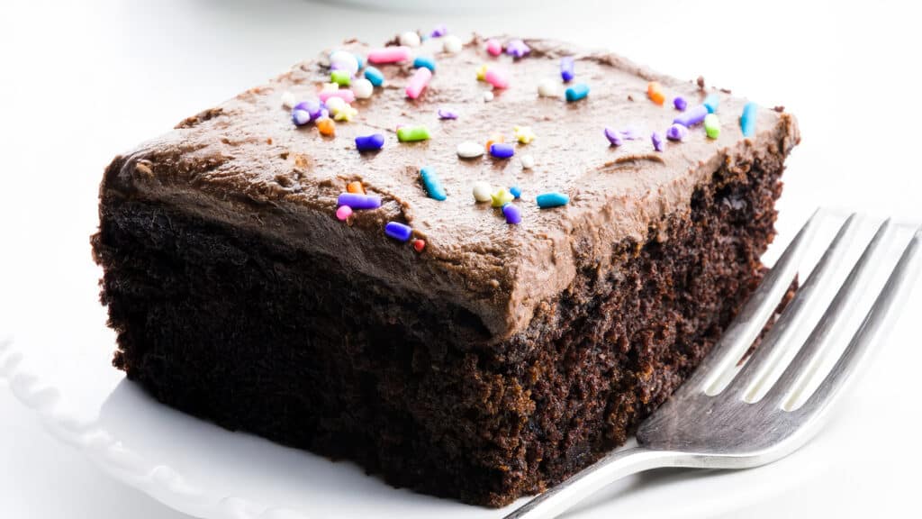 A slice of rich chocolate cake with creamy frosting, topped with colorful sprinkles, sits on a white plate. A fork rests next to the cake, ready to be used.