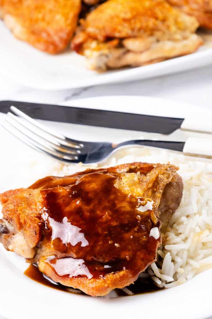 A plate with a serving of crispy chicken thighs covered in a rich, glossy bourbon glaze, placed on a bed of white rice. In the background, more cooked chicken pieces are on a white platter. A fork and knife rest beside the plate—a delicious highlight from chicken recipes.