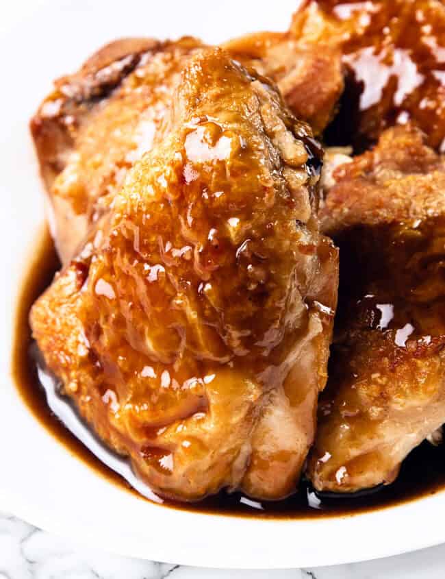 Close-up of glazed, juicy chicken pieces on a white plate. The crispy skin chicken thighs are golden-brown and covered in a shiny, thick bourbon glaze. The plate is set against a white surface with a subtle marble pattern.