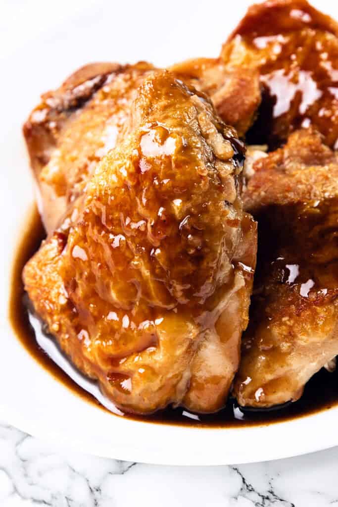 Close-up of glazed, juicy chicken pieces on a white plate. The crispy skin chicken thighs are golden-brown and covered in a shiny, thick bourbon glaze. The plate is set against a white surface with a subtle marble pattern.