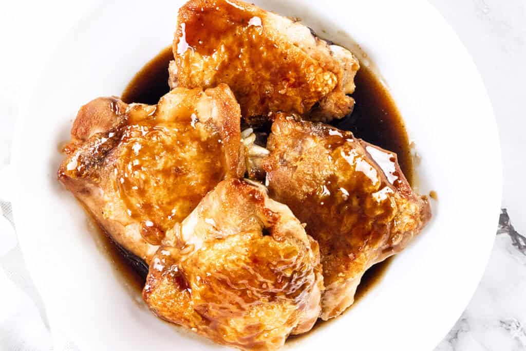 A white bowl holds four pieces of crispy skin chicken thighs, glazed to perfection. Coated in a rich bourbon glaze, the glossy sheen enhances their golden-brown allure. The light marble surface in the background complements this delicious chicken recipe.